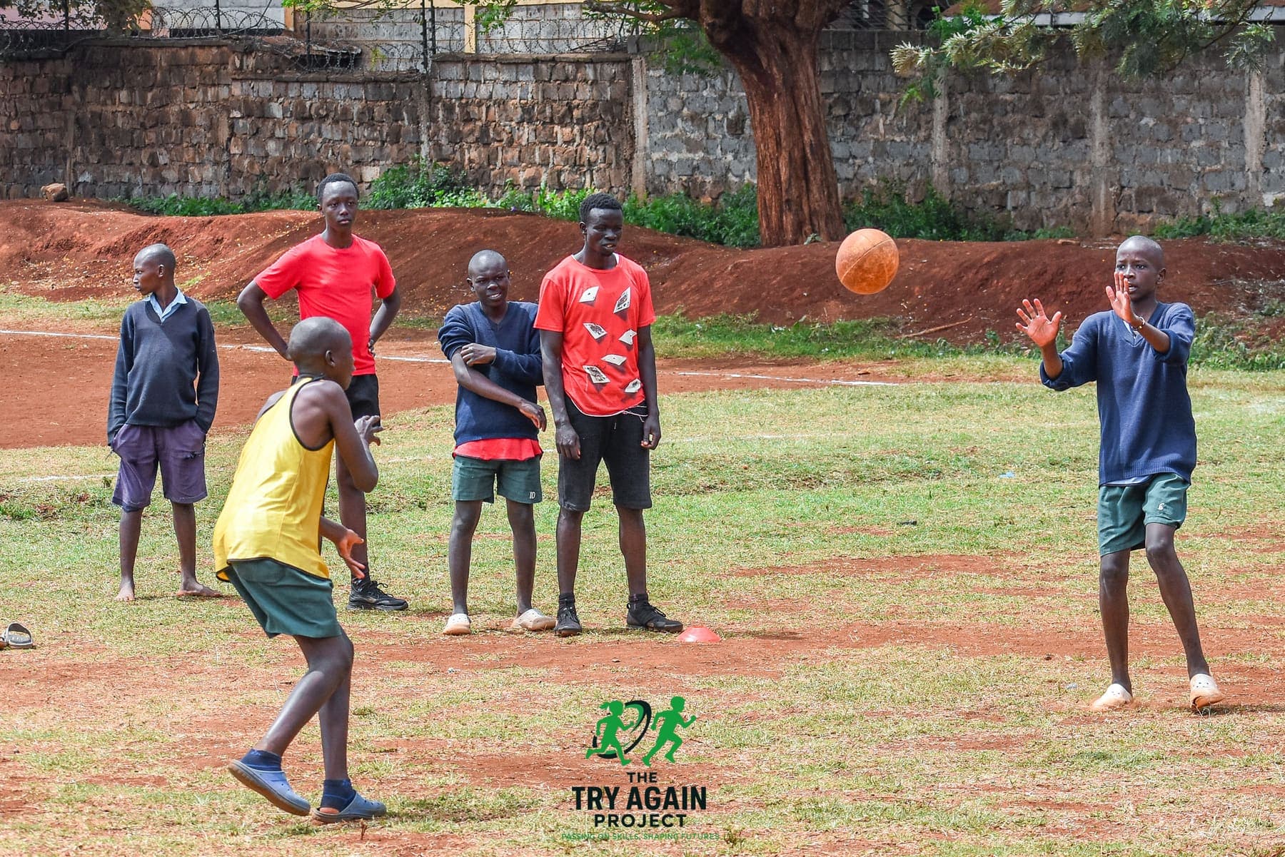 A player reaching up with both hands to take a pass as the ball drops towards him, teammates watching across the grass.