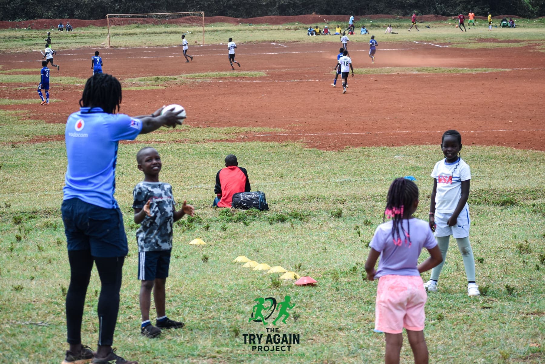 A coach passing a rugby ball to two young children on the grass while a wider session runs on the pitch behind them.