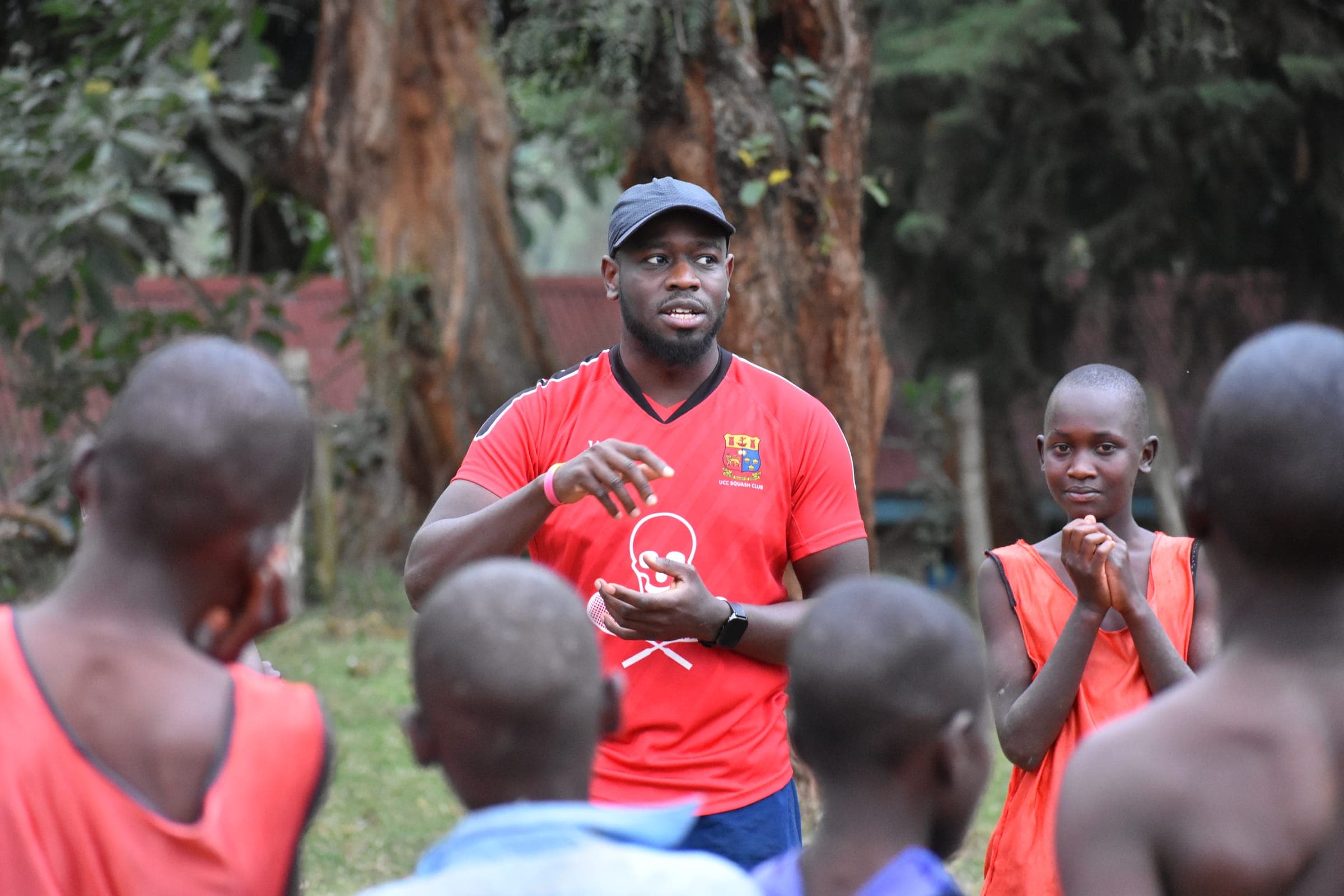A coach in a red jersey and cap talking to players gathered around him, one of them applauding.