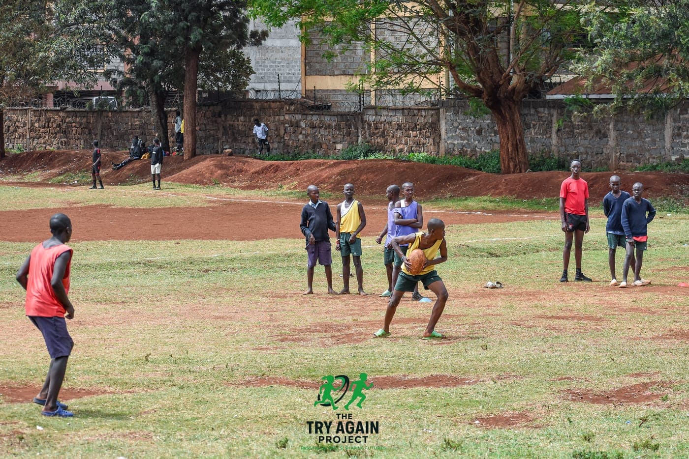 A player in a yellow bib carrying the ball forward during an open training session on a red-earth pitch, teammates spread out around him.