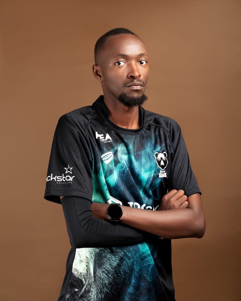 Amos Wamathi with his arms folded in a teal and black club jersey, against a plain brown backdrop.