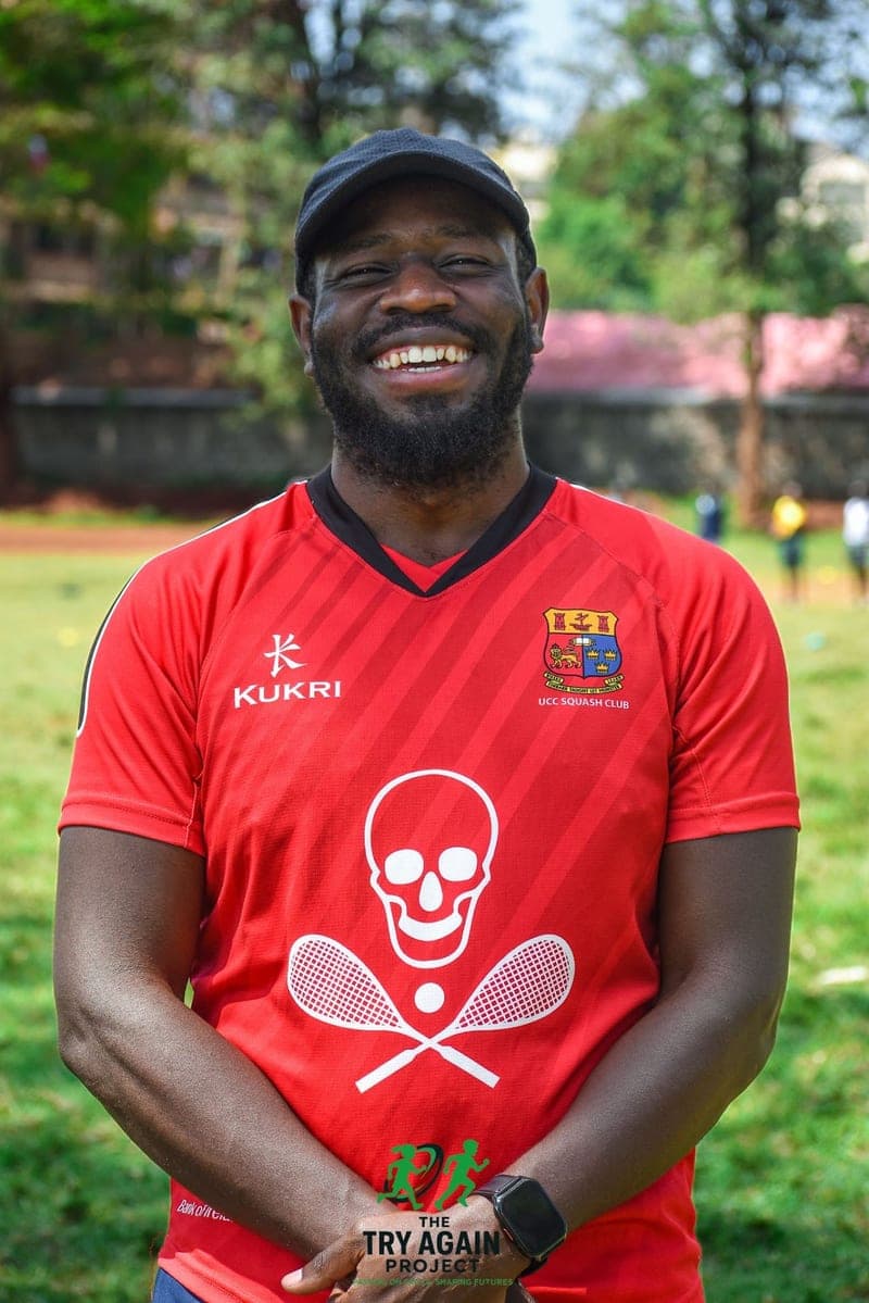 Cabral Opiyo smiling in a red training jersey and a dark cap, standing at the edge of a grass pitch.