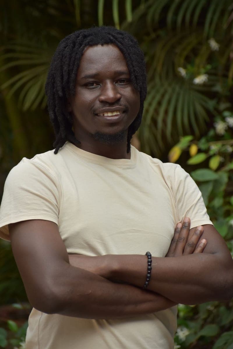 Erick Odire standing with his arms folded in a cream T-shirt, framed by green foliage.