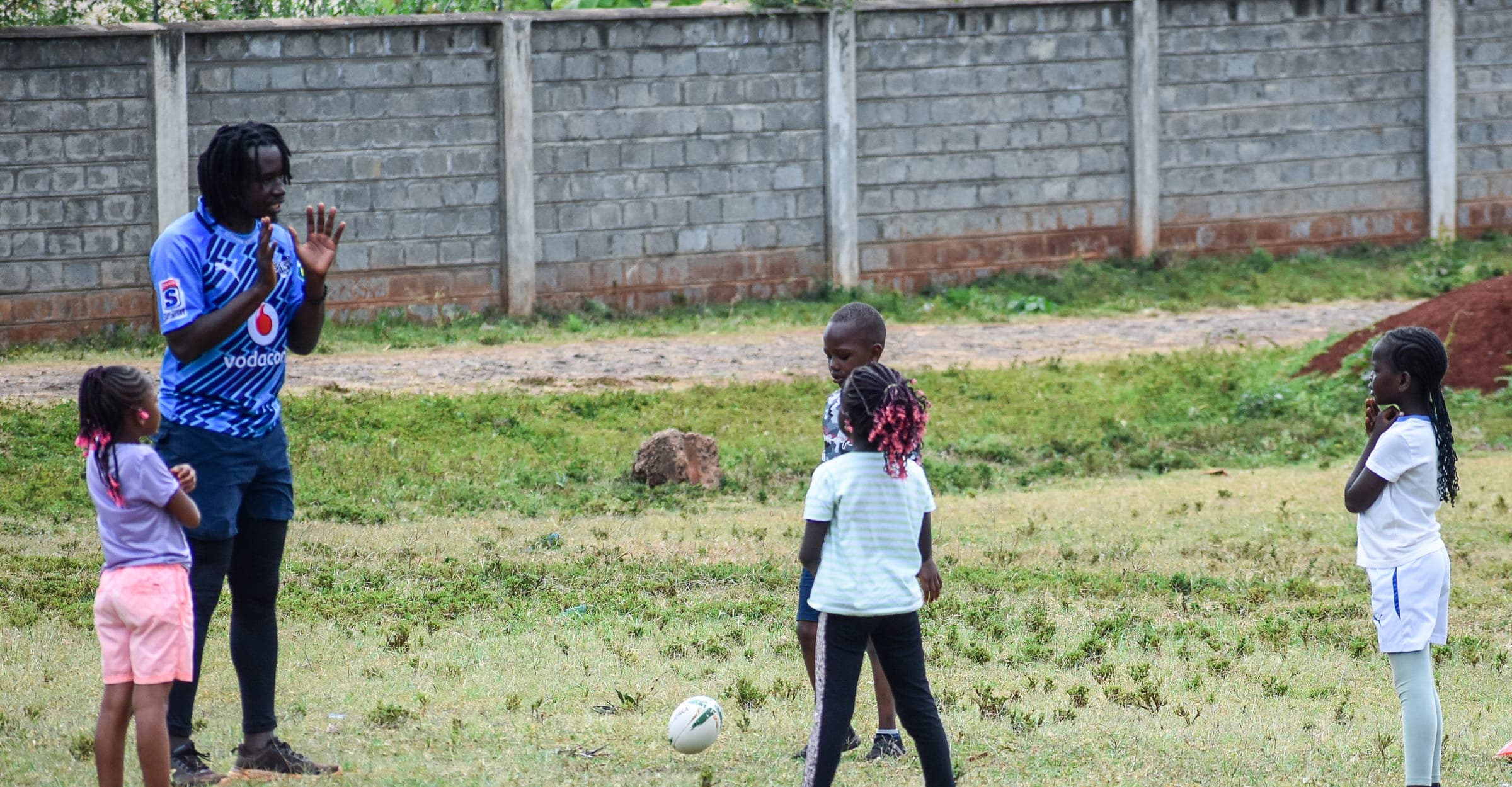 A coach working with three young girls on the grass, a rugby ball on the ground between them.